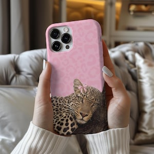 May include: A pink phone case with a leopard design and a pink bow. The top of the case has a pink leopard print. The phone is held in someone's hands.