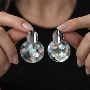 Pode incluir: Um par de brincos pendentes com uma peça superior oval em metal prateado polido ligada a uma base circular maior. A base circular é incrustada com peças de madrepérola iridescente em tons escuros dispostas num padrão de escamas sobrepostas.