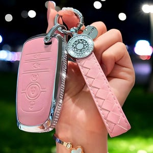 Peut inclure: Porte-clés de voiture rose avec une bordure argentée et un design incrusté de cristaux. Le porte-clés est attaché à un porte-clés en cuir tressé rose avec un charme argenté qui dit "Only Love".