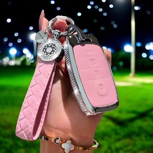 May include: A pink car key fob with a silver trim and a crystal-encrusted design. The key fob is attached to a pink braided leather keychain with a silver charm.