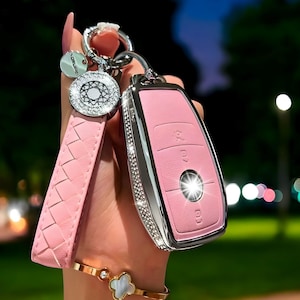 Puede incluir: Una llave de coche rosa con un borde plateado y un patrón de diamante. La llave está unida a un llavero de cuero trenzado rosa con un dije plateado.