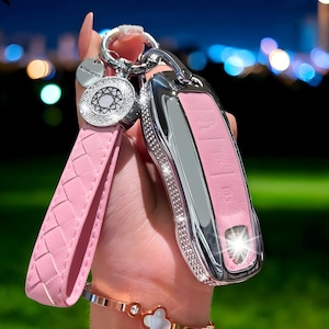 Puede incluir: Llavero de cuero rosa con una llave de coche plateada e incrustada de pedrería. El llavero tiene una correa de muñeca trenzada y un dije circular con pedrería. La llave tiene el texto "POWR FIBER".
