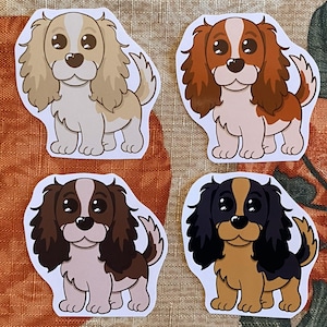 May include: Four stickers featuring cartoon Cavalier King Charles Spaniels in various color combinations: tan and white, red and white, brown and white, and black and tan. Each sticker depicts a cute dog with big eyes and a friendly expression.