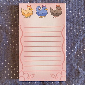 May include: A pink notepad with three cartoon chickens at the top. The chickens are yellow, blue, and brown, each wearing a hat. The notepad has horizontal lines for writing and a floral design at the bottom.
