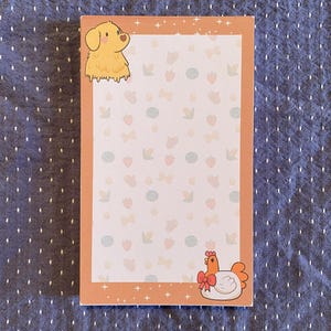 May include: A rectangular notepad with a light orange border and a patterned interior. The pattern includes strawberries, bows, and leaves. A cartoon dog and chicken are featured in the corners. The notepad is on a blue dotted surface.