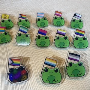 May include: A collection of green frog pins, each holding a flag with various LGBTQ+ pride flag designs. The frogs have a cartoon style with big eyes and are set against a white background.