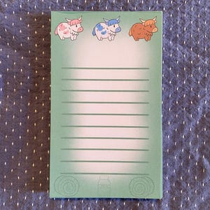 May include: A rectangular notepad with a light green background and horizontal lines. The top of the notepad features three cartoon highland cows in pink, blue, and brown. The bottom has a milk bottle illustration. The notepad is approximately 4 inches wide and 6 inches tall.