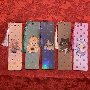 May include: Five colorful bookmarks with tassel accents. Each bookmark features a different cartoon woman illustration. The backgrounds vary, including patterns of mushrooms, strawberries, and coffee cups. The tassels are in shades of pink, green, purple, and cream.