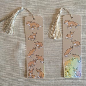 May include: Two rectangular bookmarks with tan backgrounds and illustrations of foxes in various poses. Each bookmark has a cream-colored tassel attached at the top. The foxes are in shades of orange and brown.