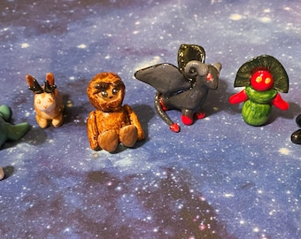 May include: A collection of handmade polymer clay figurines, including a green sea creature, a brown sloth, a black bat, a red and black dragon, a green alien, and a white ghost.