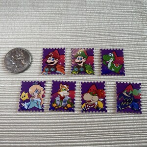 Puede incluir: Una colecci&oacute;n de siete pegatinas rectangulares moradas con varios personajes de la franquicia de videojuegos Super Mario. Las pegatinas incluyen a Mario, Luigi, Yoshi, Rosalina, Fox, Bowser y un personaje con una m&aacute;scara azul. Se incluye una moneda como referencia de tama&ntilde;o.