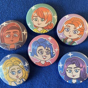 May include: A collection of six round pinback buttons featuring cartoon illustrations of women with various hairstyles and colors. Each button has a different background color, including green, pink, purple, and blue, with white star accents.