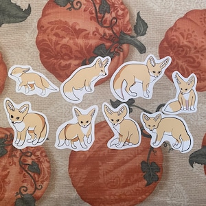 May include: A collection of eight stickers featuring illustrations of fennec foxes. The stickers depict the foxes in various poses, with tan and white fur, and large ears. The stickers are on a patterned orange and brown background.