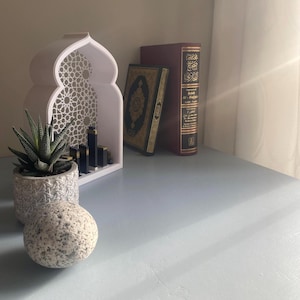 Peut inclure: Étagère décorative blanche avec un motif géométrique, présentant une petite plante succulente en pot et une pile de livres. Les livres sont reliés en bordeaux et noir avec des lettres dorées. Le texte sur le livre est "Sahih Al-Bukhari".