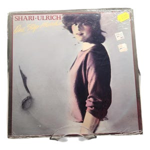 Puede incluir: Un álbum de vinilo vintage titulado "One Step Ahead" de Shari Ulrich. La portada del álbum presenta a una mujer con un suéter malva. El álbum está sellado en plástico y se muestra en un soporte de plástico transparente.