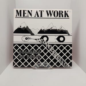 以下が含まれることがあります： 白黒のアルバムカバーで、「MEN AT WORK」と「BUSINESS AS USUAL」の文字が書かれています。カバーには、電話機とコイル状のコード、背景に山脈が描かれたスタイリッシュな画像が描かれています。