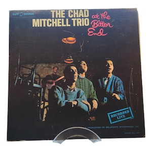 Può includere: Copertina di un album in vinile vintage per "The Chad Mitchell Trio at the Bitter End". La copertina presenta uno sfondo scuro con i membri della band in primo piano e il testo "THE CHAD at the MITCHELL TRIO Bitter End".