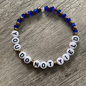 May include: A beaded bracelet with the words "YOU DO NOT YIELD" spelled out in white letter beads. The bracelet also features blue and gold beads, strung together on a clear elastic cord.