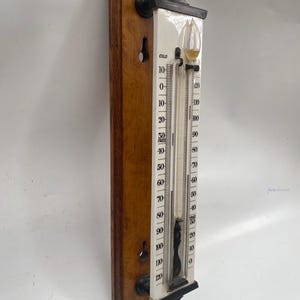 Op de afbeelding: Een antieke thermometer gemonteerd op een houten plank. De thermometer heeft een witte wijzerplaat met zwarte markeringen en een glazen buis gevuld met vloeistof. De houten plank is donkerbruin van kleur. De thermometer meet van -40 tot 120 graden Fahrenheit (ongeveer -40 tot 49 graden Celsius).