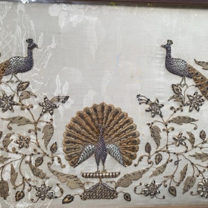 May include: A framed textile art piece featuring three embroidered peacocks on a cream-coloured fabric background. The central peacock displays a fully fanned tail, while two flanking peacocks are perched on branches surrounded by metallic floral motifs.