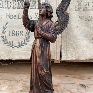 May include: A standing angel figurine with a weathered dark brown and gold finish. The figure features large, detailed wings, a long flowing robe, and one arm raised with a clenched fist. The sculpture is rendered in a classic, upright pose.