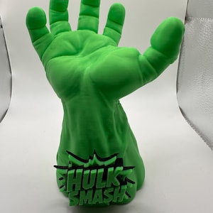 May include: A bright green, three-dimensional sculpture of a large, muscular hand with fingers spread wide. The base of the wrist features a black, jagged, comic-book-style emblem with the raised green text "HULK SMASH" centered on the front.