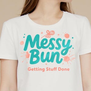 Könnte beinhalten: Weißes T-Shirt mit einer blaugrünen Grafik, auf der "Messy Bun" mit rosa Aquarellspritzer und dem Text "Getting Stuff Done" steht.