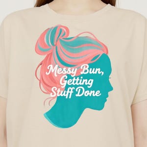 Könnte beinhalten: Ein beiges T-Shirt mit einem Grafikdruck einer Frauensilhouette mit rosa und blauen Haaren in einem unordentlichen Dutt. Der Text "Messy Bun, Getting Stuff Done" ist in Weiß auf das Shirt gedruckt.
