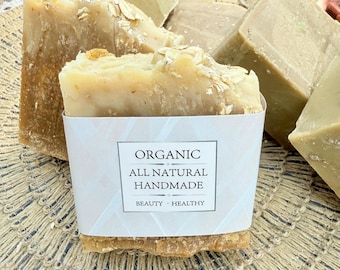 Puede incluir: Primer plano de barras de jabón orgánico hechas a mano. La barra principal está envuelta en una etiqueta blanca con las palabras "ORGANIC, ALL NATURAL, HANDMADE, BEAUTY, HEALTHY". El jabón es de color marrón claro con avena visible. Otras barras de jabón están en el fondo.