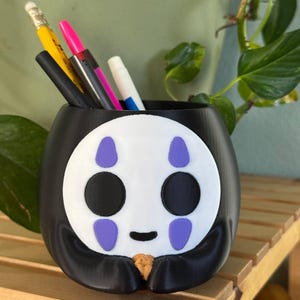 May include: Black and white No Face character from the film Spirited Away, 3D printed pencil holder. The holder has purple details and a smiling expression.