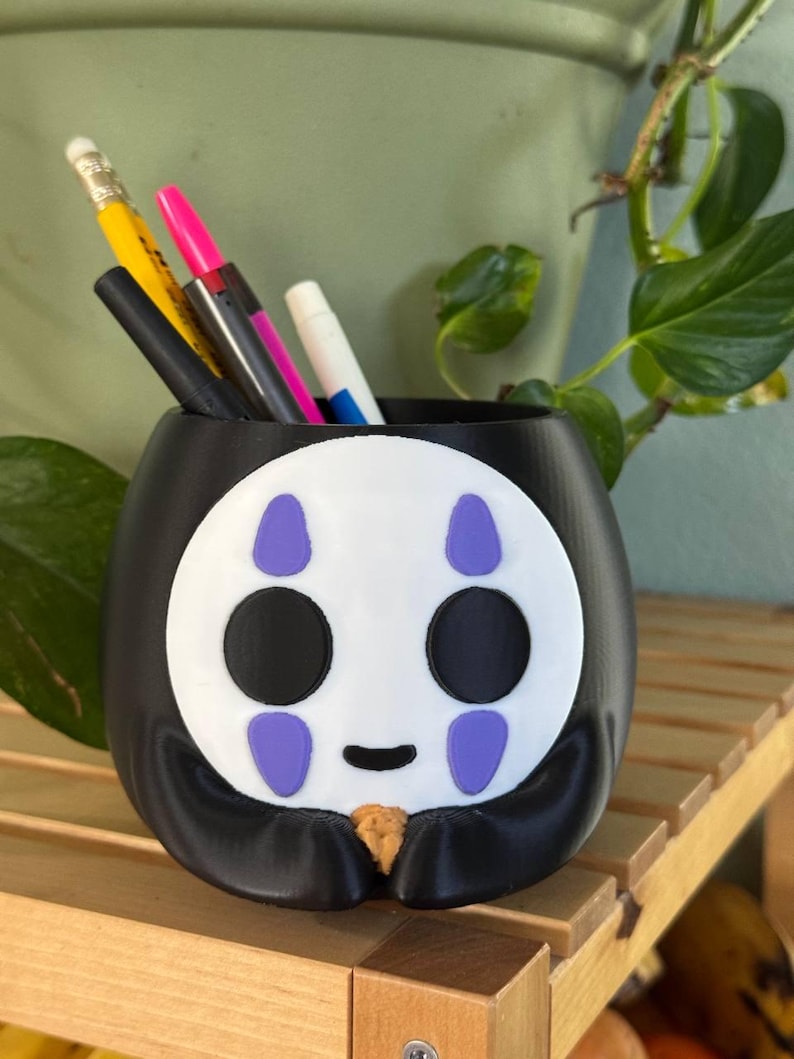 May include: Black and white No Face character from the film Spirited Away, 3D printed pencil holder. The holder has purple details and a smiling expression.