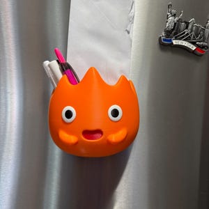 Peut inclure: Un organisateur magnétique orange en forme de flamme avec un visage de dessin animé, des yeux blancs à pupilles noires et une bouche rouge. Le récipient contient une feuille de papier blanche et trois stylos. Un aimant New York est visible.