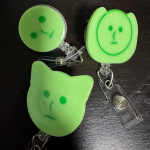 Può includere: Tre porta badge retrattili verdi con facce di cartoni animati. Uno è rotondo, uno a forma di gatto e l'altro ha una forma arrotondata con due orecchie. Ognuno ha un cinturino di plastica trasparente e una clip in metallo.