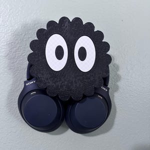 May include: A black and white 3D printed wall mount shaped like a character from the film "My Neighbor Totoro" with large white eyes. Two dark blue wireless headphones are hanging from the mount. The headphones have the word "SONY" printed on the side.