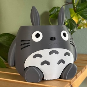 May include: A grey 3D printed planter shaped like the character Totoro from the film My Neighbour Totoro. The planter has large ears, a round body, and a small mouth.
