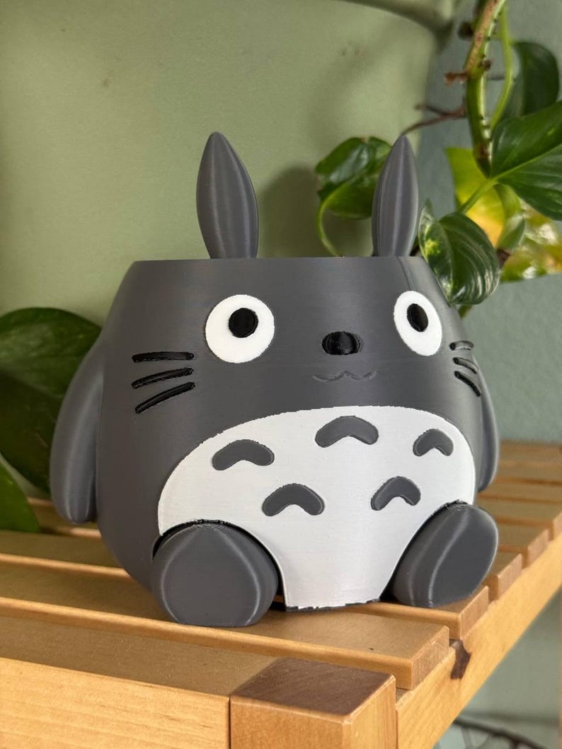 May include: A grey 3D printed planter shaped like the character Totoro from the film My Neighbour Totoro. The planter has large ears, a round body, and a small mouth.