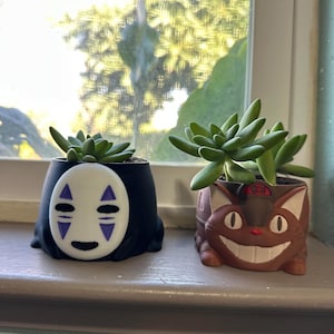 Könnte beinhalten: Zwei kleine Sukkulenten in dekorativen Keramik-Töpfen. Die Töpfe sind in Form der Figuren Ohne Gesicht und Totoro aus dem Studio-Ghibli-Film Mein Nachbar Totoro.