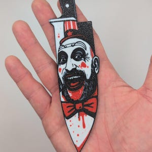 May include: A novelty patch in the shape of a knife, featuring a smiling clown face with a red bow tie and a top hat. The knife blade is white with red blood splatter and a single drop of blood.