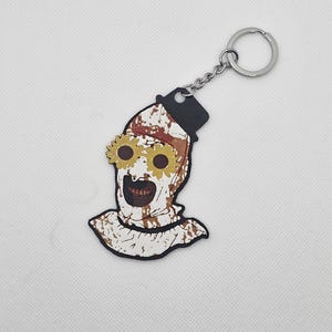 May include: A white and brown keychain featuring a character with a black top hat and sunflower eyes. The character's face is covered in blood and has a creepy smile.