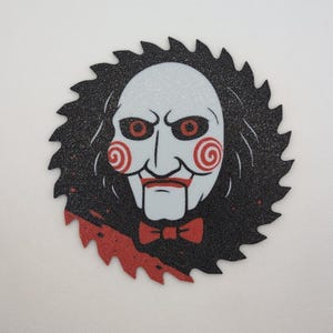 May include: A circular graphic featuring the face of the Jigsaw puppet from the Saw horror film series. The puppet's face is white with red eyes, a red mouth, and a red bow tie. The graphic is set against a black saw blade.