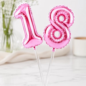 May include: A pair of metallic pink foil balloons shaped like the numbers one and eight, each attached to a clear plastic stick. The balloons feature a reflective surface and a crimped edge design, serving as a decorative cake topper.