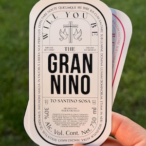 Puede incluir: Una pila de etiquetas blancas y negras con texto, incluyendo "WILL YOU BE THE GRAN NINO". Las etiquetas presentan una cruz con palomas y texto en espa&ntilde;ol. Las etiquetas tambi&eacute;n indican "750 ml" y "30% Alc."