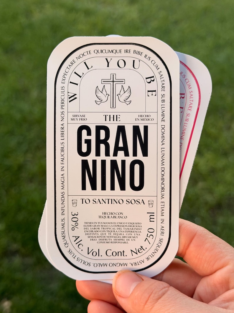 Puede incluir: Una pila de etiquetas blancas y negras con texto, incluyendo "WILL YOU BE THE GRAN NINO". Las etiquetas presentan una cruz con palomas y texto en espa&ntilde;ol. Las etiquetas tambi&eacute;n indican "750 ml" y "30% Alc."