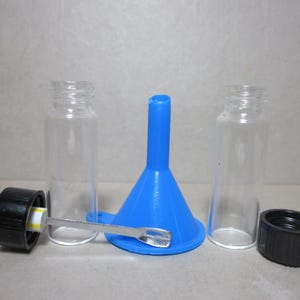 May include: A clear glass vial set with two vials, two black caps, a blue funnel, and a small silver spoon. The vials are cylindrical with threaded openings. The funnel is cone-shaped.