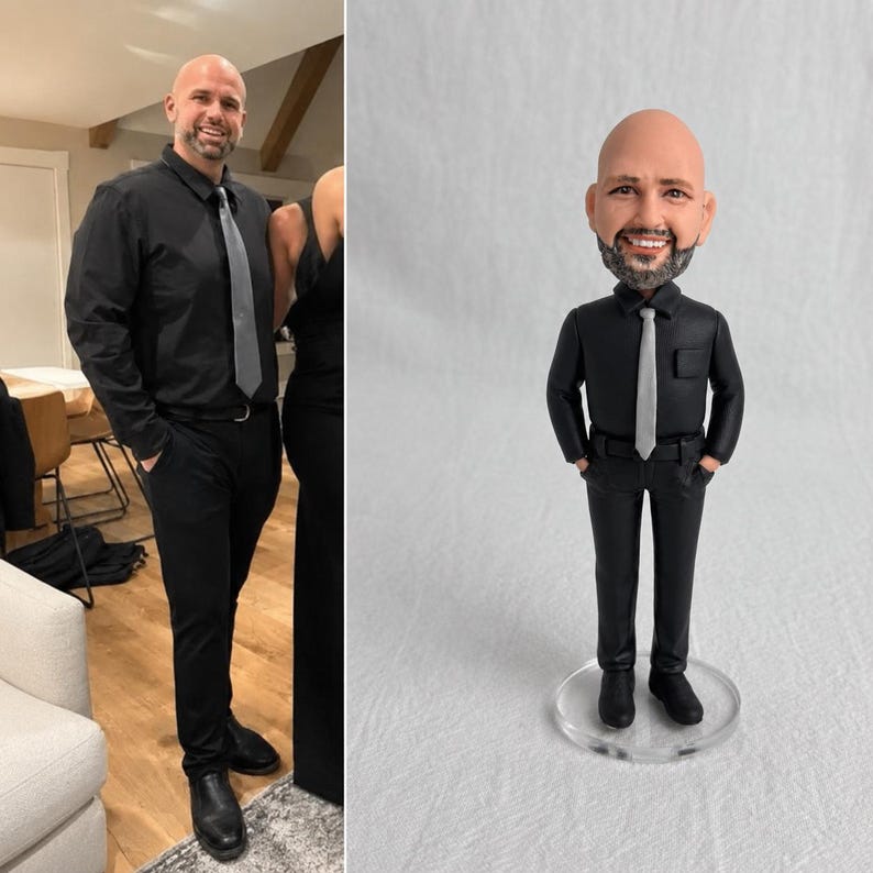 May include: A custom bobblehead figurine depicting a bald person with a beard, wearing a black long-sleeved button-down shirt, a light gray necktie, and black trousers. The figure stands on a clear circular base with hands in pockets.
