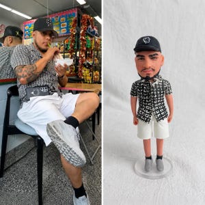 May include: A custom figurine depicting a person wearing a black baseball cap with a white clover logo, a black and white patterned short-sleeve button-down shirt, white shorts, black socks, and grey sneakers, standing on a clear circular base.