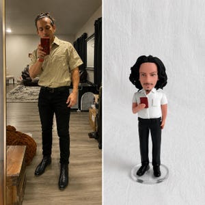 May include: A custom bobblehead figurine featuring a person with dark wavy hair and a goatee, wearing a white short-sleeved collared shirt, black trousers, and black shoes. The figure holds a small red rectangular object in the right hand.