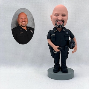 May include: A custom bobblehead figurine of a police officer in a dark uniform, holding a helmet and handcuffs. The figurine is on a grey base. An oval portrait of the person is to the left. The bobblehead has a smiling face and a black beard.