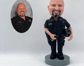 May include: A custom bobblehead figurine of a police officer in a dark uniform, holding a helmet and handcuffs. The figurine is on a gray base. An oval portrait of the person is to the left. The bobblehead has a smiling face and a black beard.