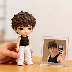 May include: A stylized figurine with brown curly hair, wearing a black tank top, white pants, and white sneakers, standing next to a clear acrylic frame containing a photograph of a person with similar hair wearing a black tank top and a necklace.